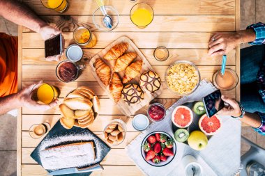 Kahvaltı yiyecekleriyle dolu masanın dikey görüntüsü ve cep telefonuyla yiyip eğlenen bir grup insan. Aile ve arkadaşlar birlikte yemek yer ve sabah keyfine bakarlar. Fırın ve meyve.