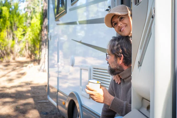 Young couple of tourists with camper motor home sitting at the door enjoying nature outdoors leisure feeling. Travel people and mountains destination concept. Man and woman living van life together