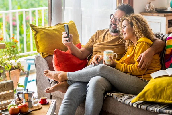 Caucasian couple doing video call with mobile phone at home sitting on the sofa. Man and woman enjoying technology wireless connection during morning breakfast and have fun calling friends