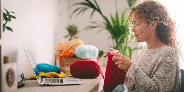 Woman at home in leisure activity learning from online class to knit work. Female people using laptop for her hobby or alternative work. Middle age lady knitting with red wool. Concept of using web