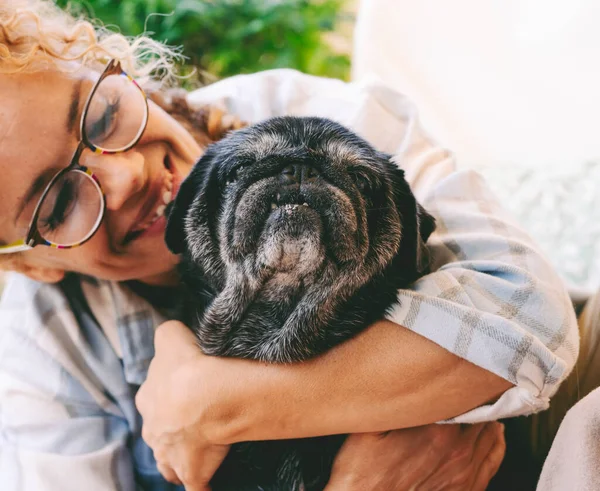 Cheerful woman hugging black old dog with love and friendship. Animal pug best friend forever. Female people and her puppy enjoying time together with happiness. Lady smiling and bonding dog