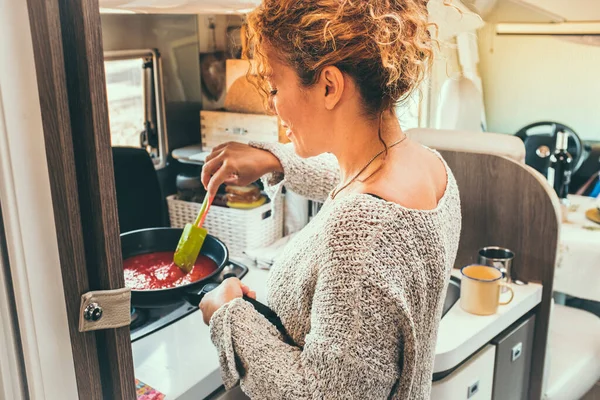 Woman viewed from back preparing and cooking tomato sauce for lunch inside a modern camper van. Off grid and van life lifestyle with traveler people having food preparation. Indoor kitchen activity