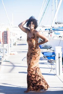 Açık havada turistik aktiviteler yapan zarif elbiseli siyahi genç bir kadının portresi. Etnik ve güzel bir kız, arka planda mavi gökyüzü olan limanda güneşlenmenin ve tatilin tadını çıkarıyor.