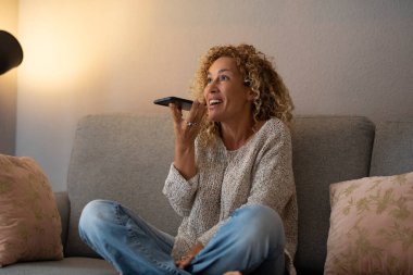Evdeki telefon görüşmesinde telefonda konuşan heyecanlı bir kadın kanepede oturuyor. Kapalı alan aktivitesi ve modern cihaz kullanımı. Dişi insanlar arkadaşlarıyla ses bağlantısından hoşlanırlar. Akıllı telefon.