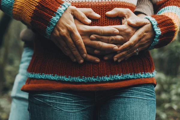 Concept of love and relationship with close up of a heterosexual mature couple hugging from back and holding hands on belly. Man and woman people embracing and holding with tenderness sweetness outdoor