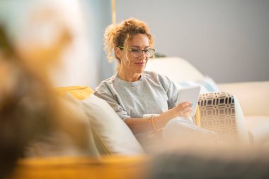 Young mature woman smile and enjoy time reading an electronic book on tablet reader device comfortably sitting on the sofa at home in indoor leisure activity alone. Female people have relaxed indoor