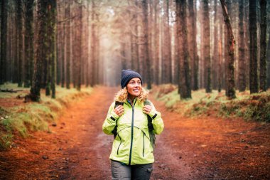 Parkta gezintiye çıkmış sırt çantalı genç bir kadın var. Doğanın tadını çıkaran, gülümseyen ve yürüyüş yapan bir kadın. Arka planda ağaçlar. Orman manzaralı bir yer.