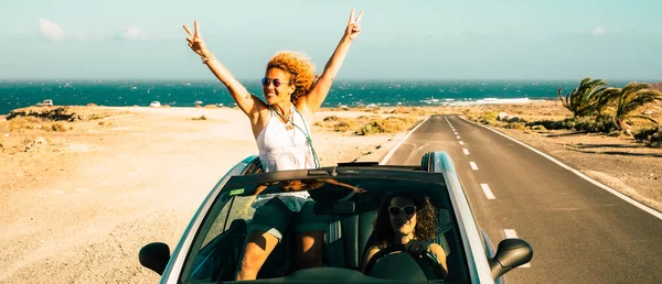 Travel with car vehicle and tourists summer destination concept. Happy and excited couple of women friends drive and have fun with a convertible and long straight road in background. Ocean and sea