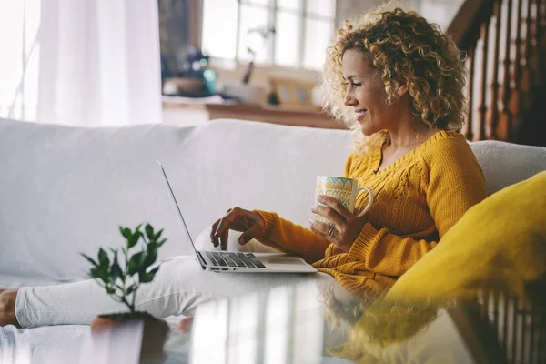 Real life of woman using laptop computer smiling and drinking a coffee at home comfortably sitting on the sofa. Modern female people use computer notebook in indoor leisure activity. Technology
