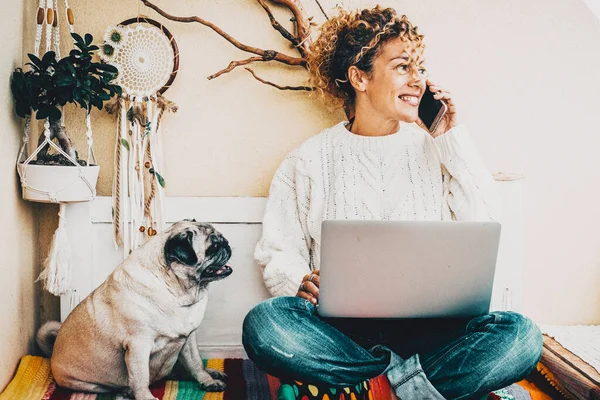 Woman smiles and talks at the phone with her best friend dog sit down near her. Modern people smart working lifestyle using computer and enjoying animal friendship with a pug. Concept of couple