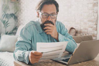 One man at home reading worried  electricity or costs bill in front of a laptop. People at home with economy problems. Online payment house cost. One man working on a computer inside the apartment. Job