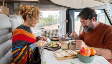 Young mature couple enjoy time together having lunch inside camper van. Happy people in free leisure holiday vacation with vehicle transport lifestyle. Alternative off grid house and freedom day