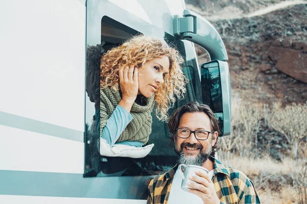 Two people, man and woman enjoying travel road trip destination together. Female looking outside her camper van and bearded male drinking coffee outdoor. Off grid alternative house concept. Van life