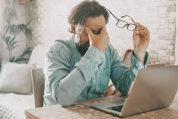 Tired man holding glass and touching is eyes after over work on notebook computer. Online activity and problems to solve concept. Modern worker people using laptop at table desk. Economy difficulty