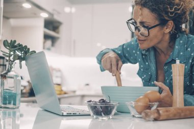 Modern wife prepare breakfast or lunch using computer and online tutorial in the kitchen at home. Real life woman cooking alone. Female people cook happy watching laptop and smiling. House lifestyle