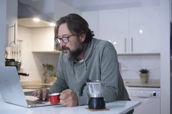 man at home having breakfast before work, drinking Italian coffee. Modern people using computer laptop in the kitchen in house real lifestyle leisure. Male person indoor online activity at home