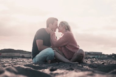 Kız ve oğlanla romantik bir akşam kumsalda oturup şefkatle öpüşüyorlar. İki insan, kadın ve erkek, kumda oturmuş günbatımının tadını çıkarıyorlar. Erkek kadın öpücüğü