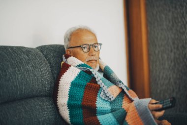 Yaşlı bir adam, düşük sıcaklık ve soğuk algınlığı belirtileri için renkli yün örtüsüyle evdeki kanepede oturuyor. Enerji gazı krizi konsepti. İçerideki ısıdan tasarruf ediyoruz. Kışın insan kavramı