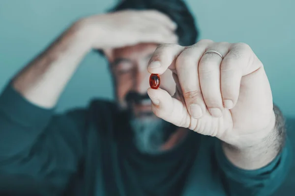 Close up of man holding red pill against migraine headache. Person in background holding and touching his front with pain. Concept of pharmacy and medicine treatment. Focus on hand with pills