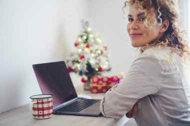 Çekici, genç, yetişkin bir kadının önünde dizüstü bilgisayarla gülümseyen ve düşünen portresi ve arka planda Noel süslemeleri. Mutlu kadınlar evlerinde Noel tatili aktivitelerinin keyfini çıkarırlar.