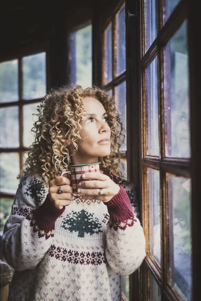 Pensive cute woman alone at home looking outside the window and drink cup of tea or coffee alone. Christmas holiday season. Young female people wearing xmas sweater and enjoy december season evening