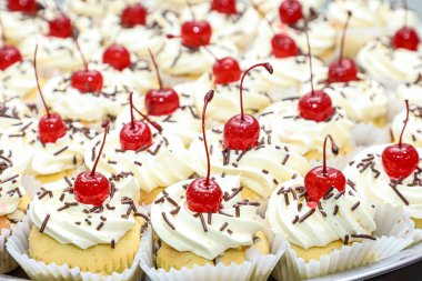 Various mini cakes with whipped cream, chocolate sprinkles and cherries on top, as the focus on these.