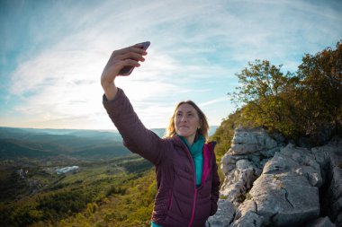 Kız gün batımında dağın tepesinde selfie çekiyor. Dağları dolaşın. Kız fotoğraf çekiyor.