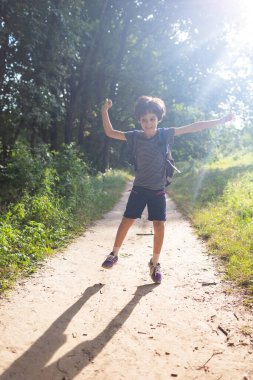 Bir okul çocuğu parkta yürüyor, sırt çantalı bir çocuk orman yolunda zıplıyor, bir çocuk doğada eğleniyor