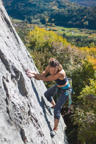 女攀岩者在秋天的森林和蓝天的背景下爬上岩石 一个登山者用设备走了一条困难的路 — 图库照片