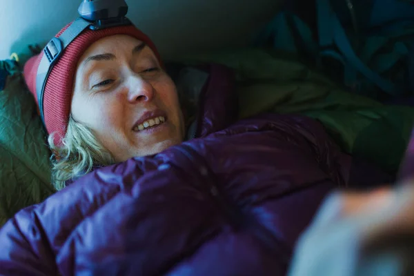 Woman in a sleeping bag in a tent, winter trekking, a woman with a headlamp lies in a sleeping bag in a tent and holds a smartphone