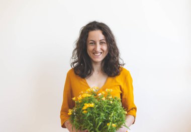 Bir kadın ve çiçekli bir saksı, sarı elbiseli gülümseyen bir kız beyaz bir duvarın arka planında oturuyor, iç mekan bitkisi