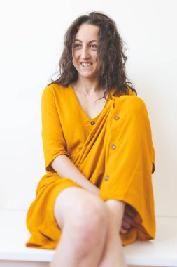 Sarı elbiseli gülümseyen kız beyaz bir duvarın arkasında oturuyor, kıvırcık saçlı bir kadının portresi.