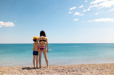 Anne ve çocuk sahilde durur ve denize bakarlar. Deniz kenarında tatiller.