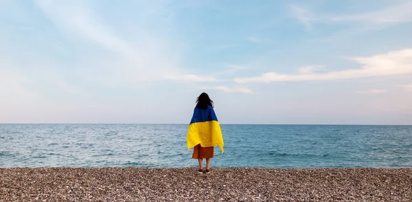 A girl stands by the sea with a Ukrainian flag, support for Ukraine, a sad Ukrainian woman, the flag flutters in the wind