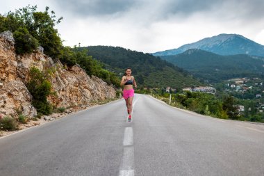 Bir kız asfalt bir yolda koşuyor güzel dağların arka planında, bir kadın akşam koşusunda, aktif bir yaşam tarzı, ince bir esmer dağ yolunda koşuyor.