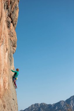Bir kadın mavi gökyüzüne karşı bir kayaya tırmanır, güçlü bir kız gücü ve dayanıklılığı eğitir, ekstrem bir spor, doğal zemine kaya tırmanışı, bir kaya tırmanışçısı iple tırmanır.