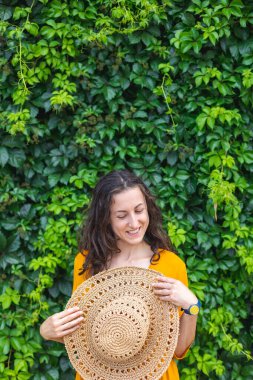 Hasır şapkalı gülümseyen bir kız sarmaşıkla kaplanmış bir duvarın yanında duruyor. Siyah saçlı bir kadının portresi.