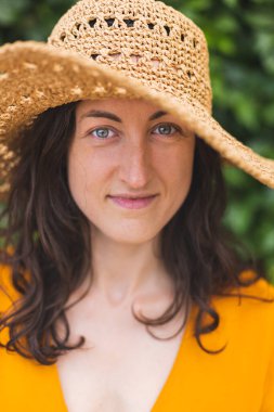 Hasır şapkalı gülümseyen bir kız sarmaşıkla kaplanmış bir duvarın yanında duruyor. Siyah saçlı bir kadının portresi.