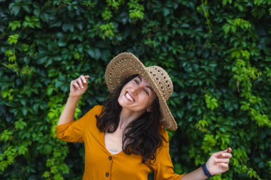 Hasır şapkalı gülümseyen bir kız sarmaşıkla kaplanmış bir duvarın yanında duruyor. Siyah saçlı bir kadının portresi.