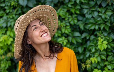 Hasır şapkalı gülümseyen bir kız sarmaşıkla kaplanmış bir duvarın yanında duruyor. Siyah saçlı bir kadının portresi.