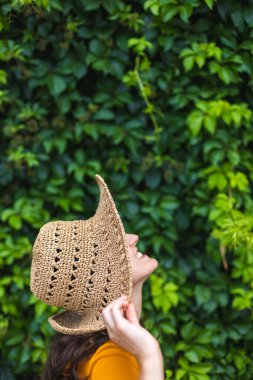 Hasır şapkalı gülümseyen bir kız sarmaşıkla kaplanmış bir duvarın yanında duruyor. Siyah saçlı bir kadının portresi.