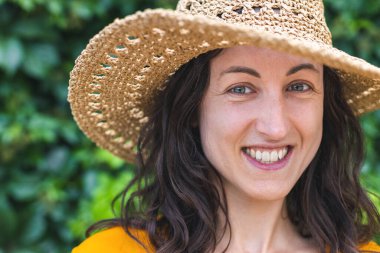 Hasır şapkalı gülümseyen bir kız sarmaşıkla kaplanmış bir duvarın yanında duruyor. Siyah saçlı bir kadının portresi.