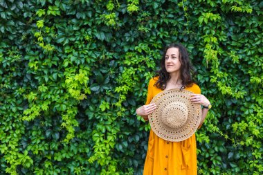 Hasır şapkalı gülümseyen bir kız sarmaşıkla kaplanmış bir duvarın yanında duruyor. Siyah saçlı bir kadının portresi.