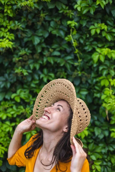 Hasır şapkalı gülümseyen bir kız sarmaşıkla kaplanmış bir duvarın yanında duruyor. Siyah saçlı bir kadının portresi.