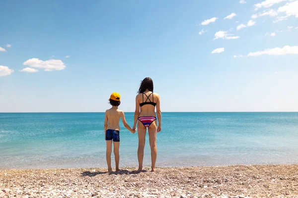 Anne ve çocuk sahilde durur ve denize bakarlar. Deniz kenarında tatiller.