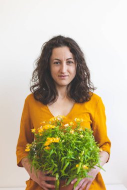 Bir kadın ve çiçekli bir saksı, sarı elbiseli gülümseyen bir kız beyaz bir duvarın arka planında oturuyor, iç mekan bitkisi