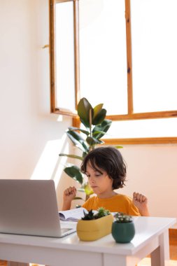Uzaklık Eğitimi, okul çocuğunun portresi, öğretmenle video görüşmesi, çocuk masaya oturur ve parmaklar, jestler için ısınır, çocuk dizüstü bilgisayara bakar.