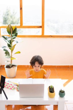 Uzaklık Eğitimi, okul çocuğunun portresi, öğretmenle video görüşmesi, çocuk masaya oturur ve parmaklar, jestler için ısınır, çocuk dizüstü bilgisayara bakar.
