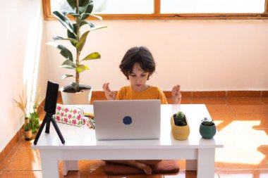 Uzaklık Eğitimi, okul çocuğunun portresi, öğretmenle video görüşmesi, çocuk masaya oturur ve parmaklar, jestler için ısınır, çocuk dizüstü bilgisayara bakar.