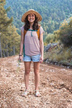 Ormanda sırt çantası ve hasır şapkalı bir kadının portresi, yürüyüş yapan gülümseyen bir kız, yaz yürüyüşü yapan, dağ yolunda yürümekten zevk alan mutlu bir kadın.